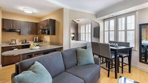 A modern kitchen with dark cabinets, a microwave, and a coffee maker. There is a dining area with a table and chairs, and a sofa with cushions.