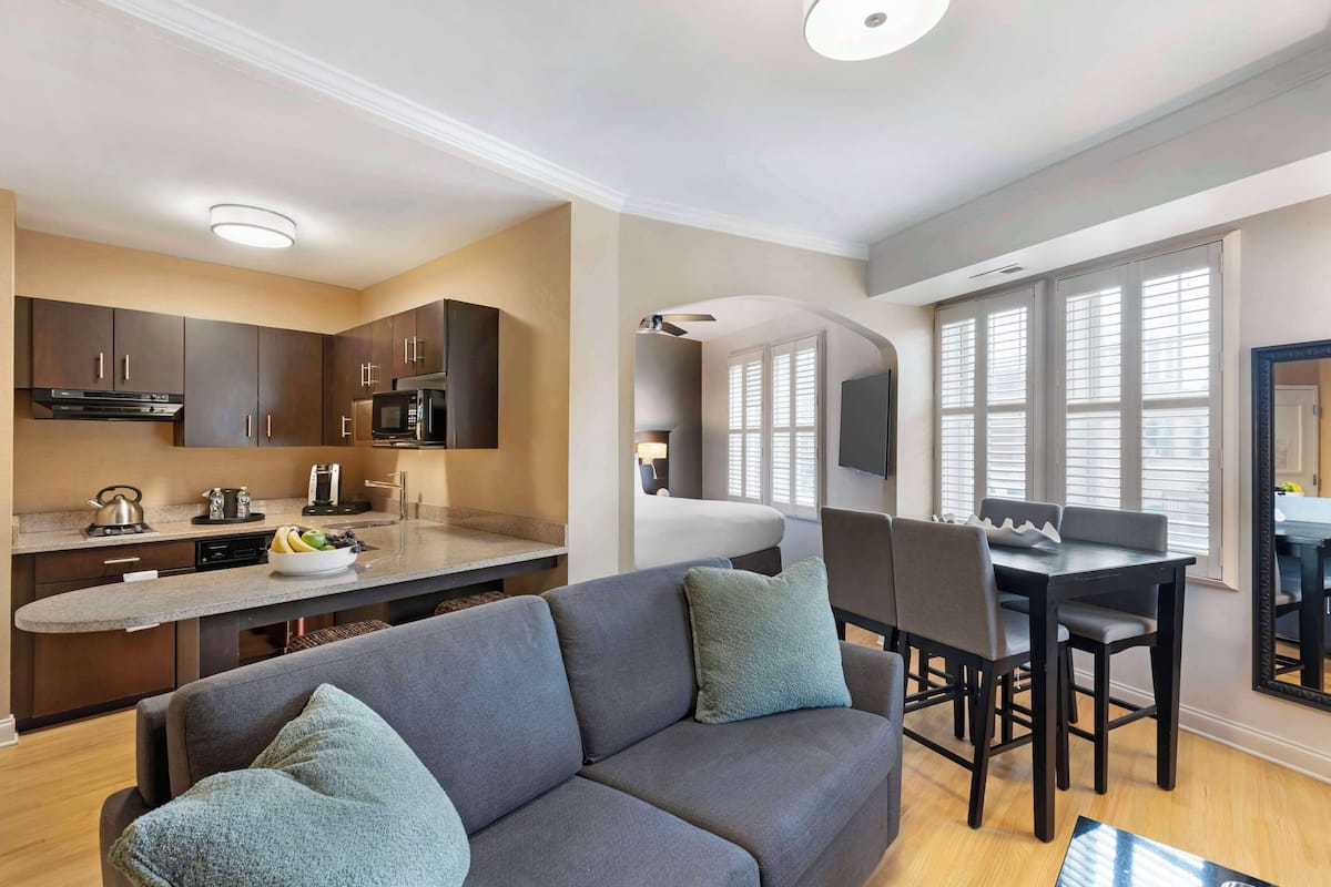 A modern kitchen with dark cabinets, a microwave, and a coffee maker. There is a dining area with a table and chairs, and a sofa with cushions.
