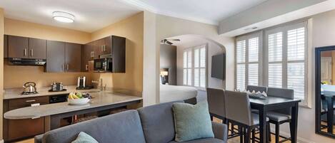 A modern kitchen with dark cabinets, a microwave, and a coffee maker. There is a dining area with a table and chairs, and a sofa with cushions.