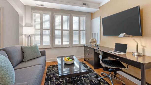 A modern hotel room with a sofa, a desk with a laptop, a large flat-screen TV, and a window with blinds. - Best Western Plus Hawthorne Terrace Hotel (Chicago)