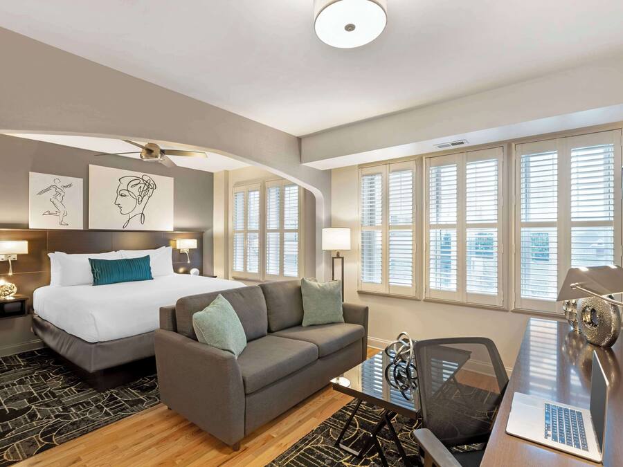 A modern hotel room with a bed, a sofa, a desk with a laptop, and large windows with blinds.