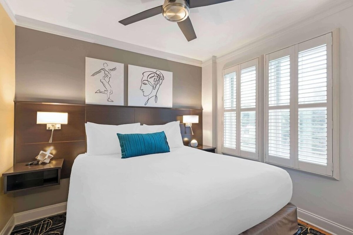 A hotel room with a large bed, two wall-mounted lamps, a ceiling fan, and artwork above the bed.