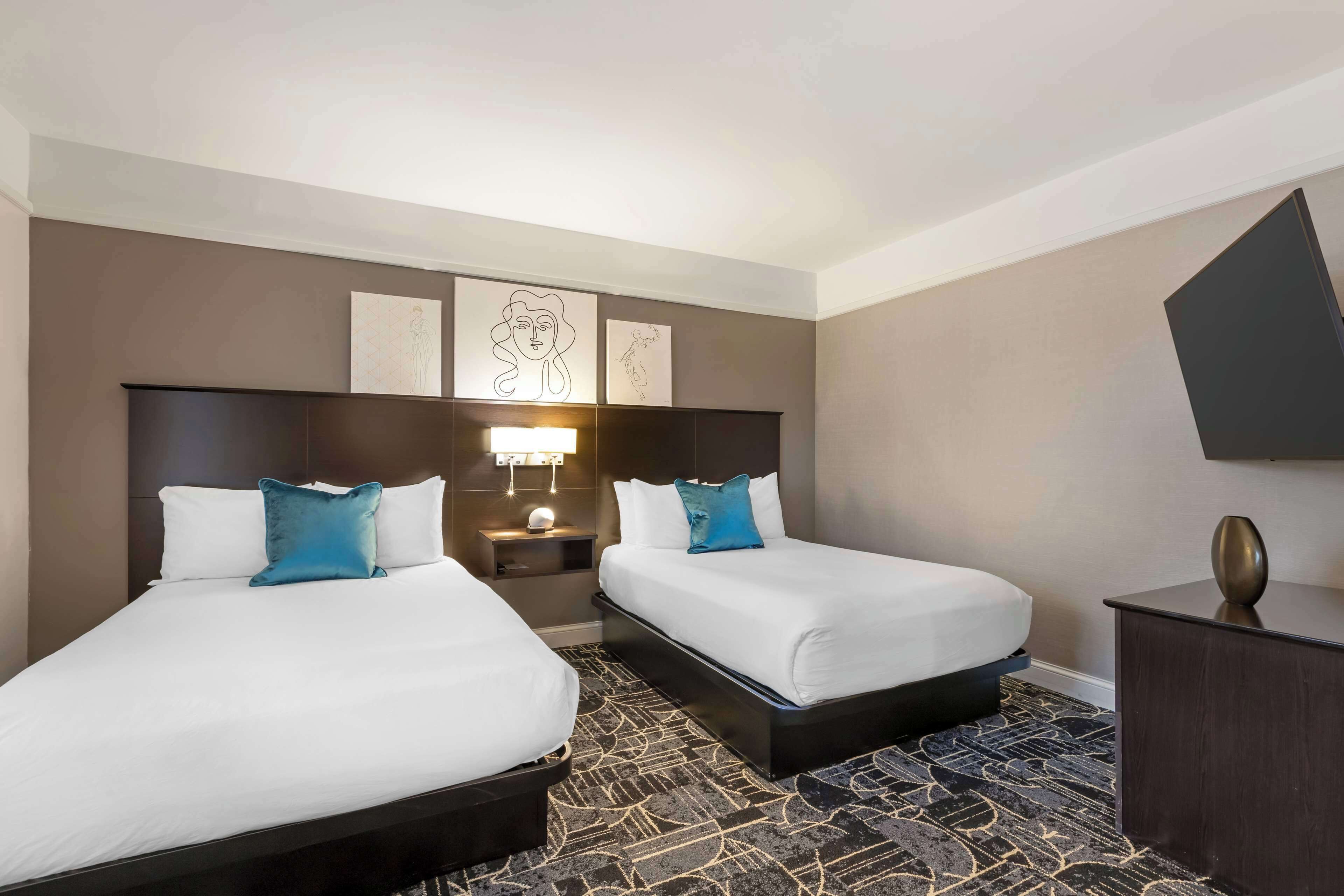A hotel room with two beds, a nightstand, a lamp, and artwork on the wall.