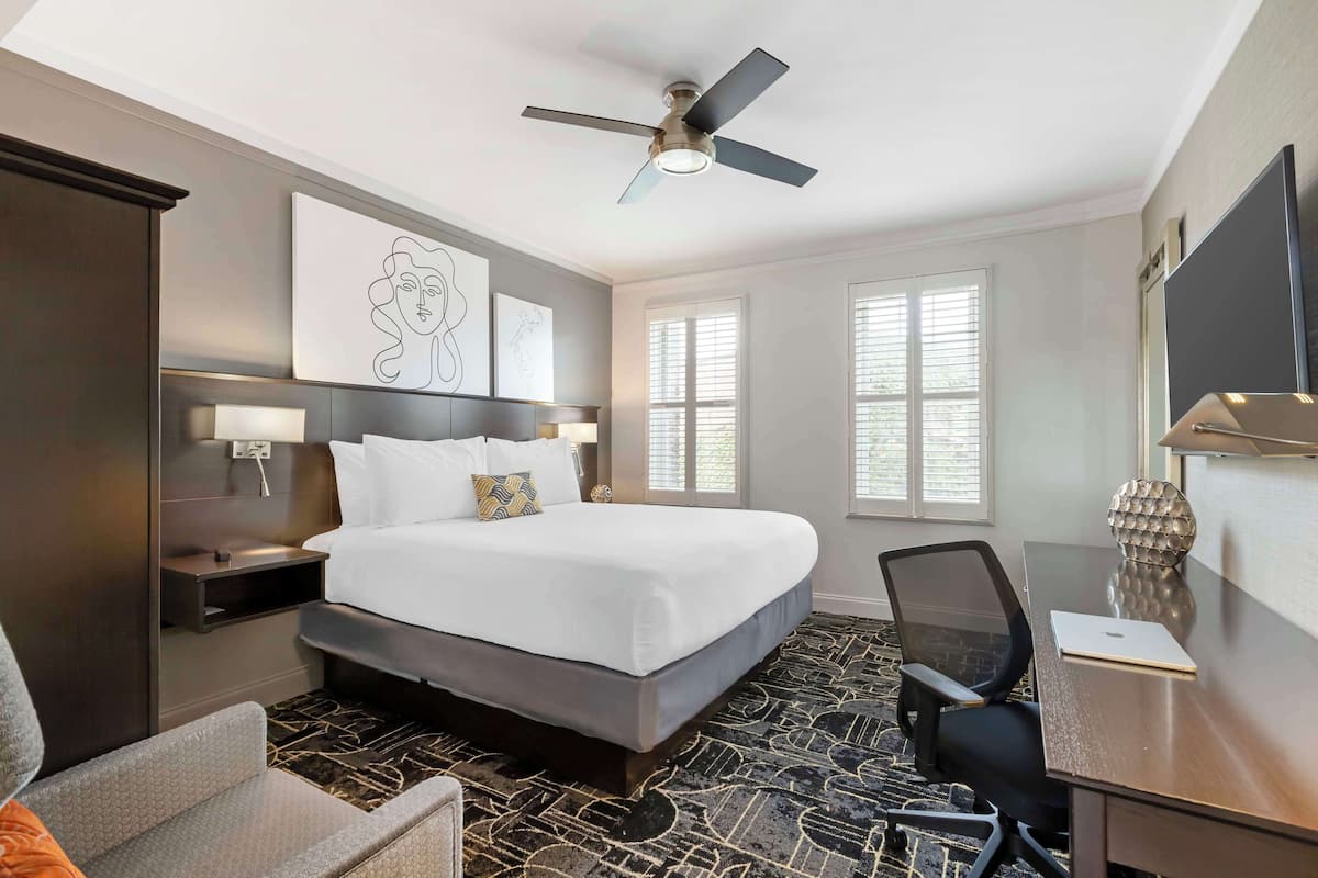 A hotel room with a large bed, a desk with a laptop, a chair, a ceiling fan, and artwork on the walls.