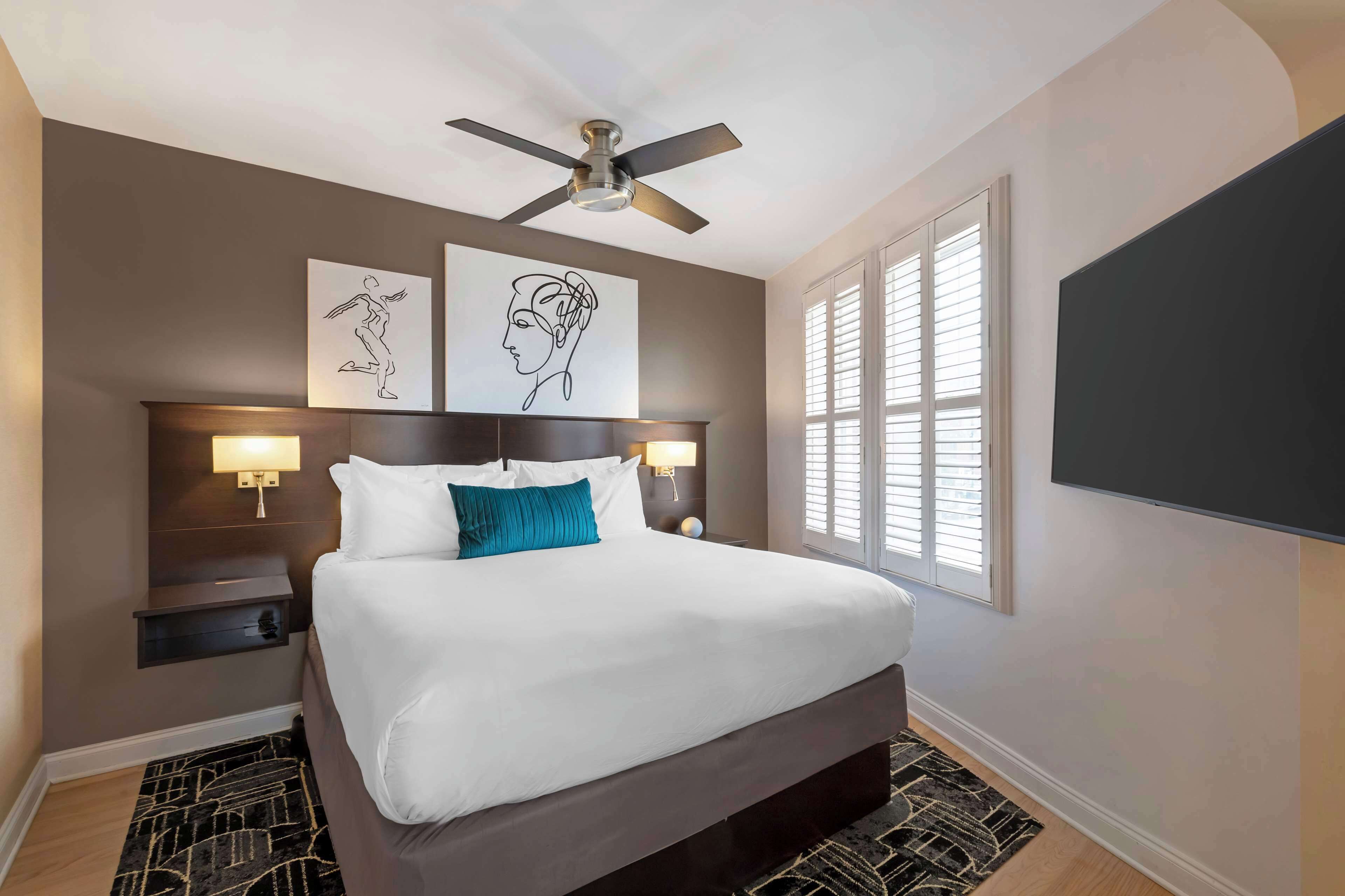 A modern hotel room with a large bed, a ceiling fan, artwork on the wall, and a TV mounted on the wall.