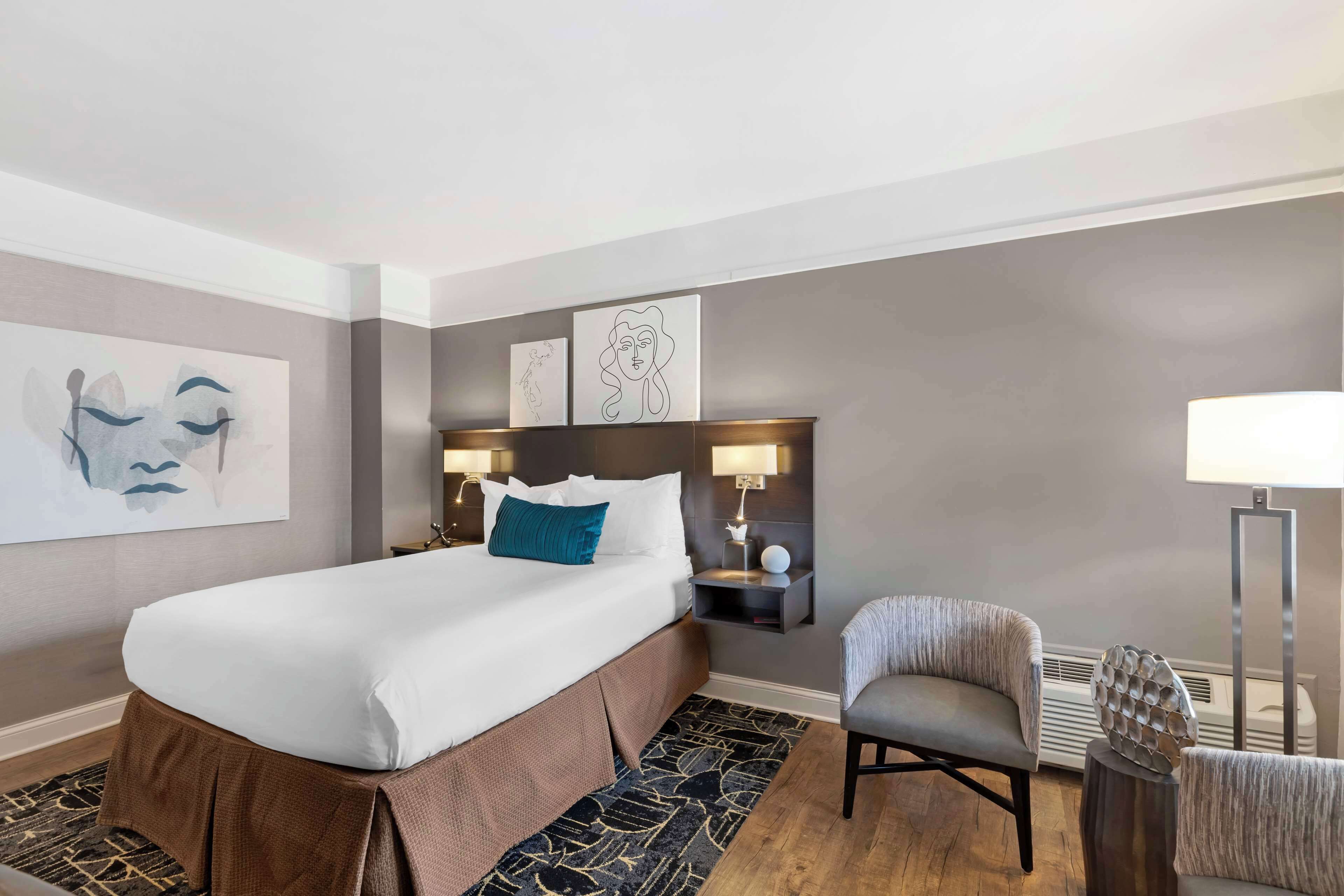 A modern hotel room with a large bed, a chair, a nightstand with a lamp, and artwork on the walls.