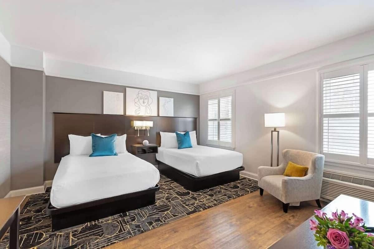 A hotel room with two beds, a chair, a lamp, and artwork on the wall.