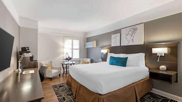 Una habitación de hotel con una cama grande, un escritorio con una computadora portátil, una silla, una mesita con una lámpara y obras de arte en las paredes.