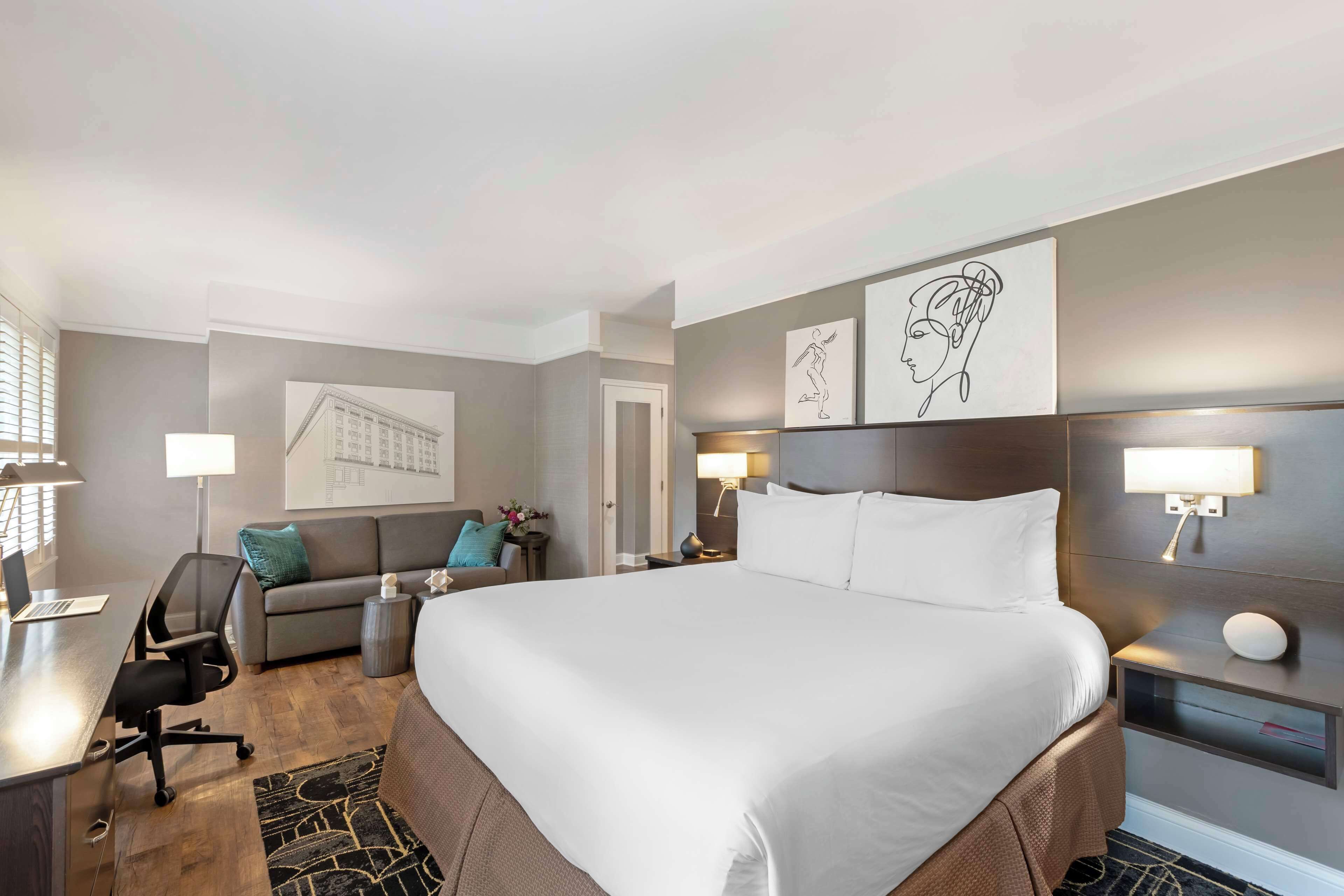 Habitación de hotel con una cama grande, un escritorio con silla, un sofá y una mesita de noche con lámpara.
