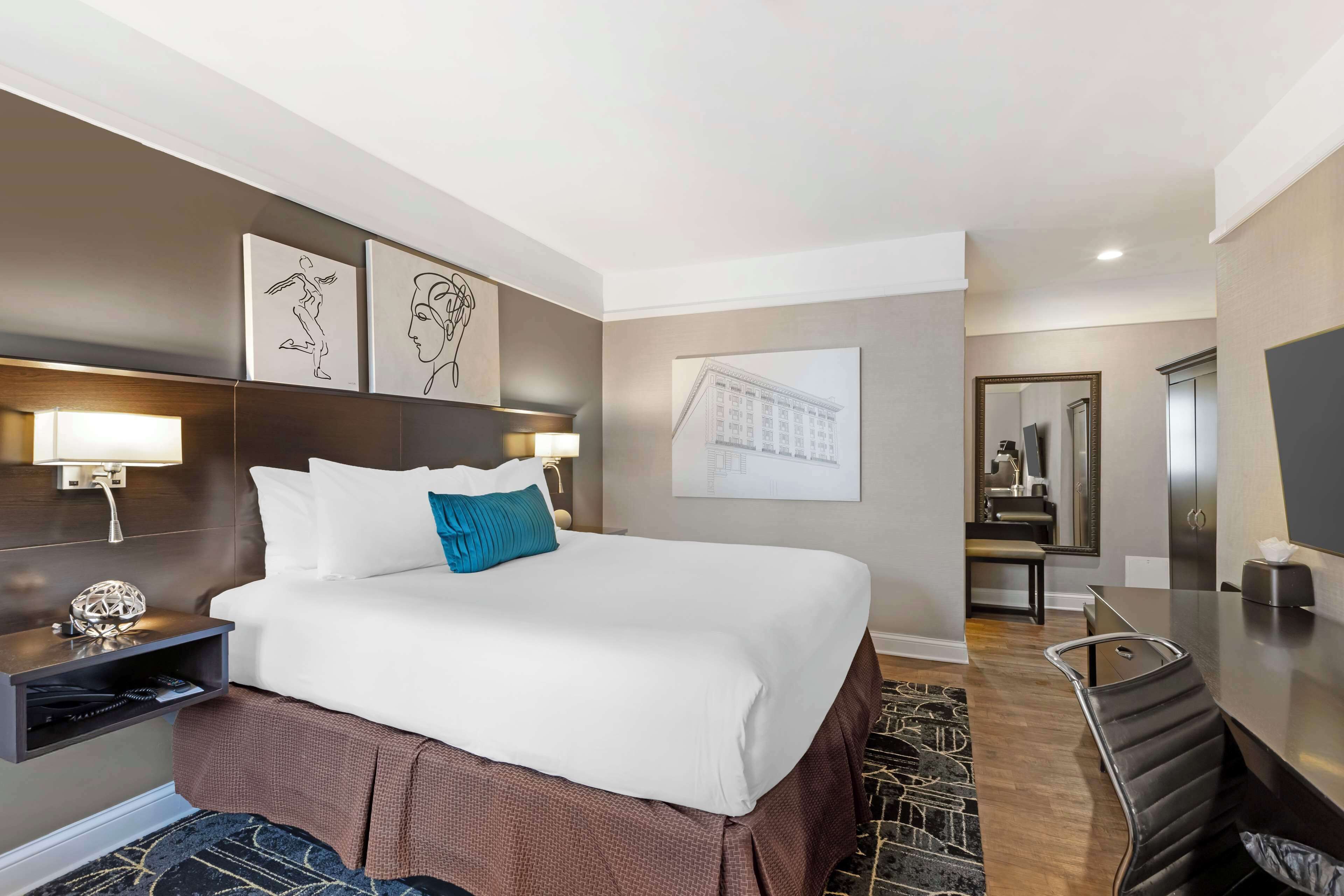 A hotel room with a large bed, a desk, a chair, and a mirror. There are wall-mounted lights and artwork above the bed.