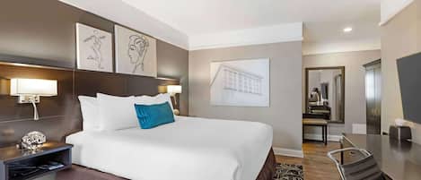 A hotel room with a large bed, a desk, a chair, and a mirror. There are wall-mounted lights and artwork above the bed.