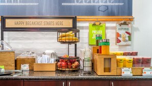 A breakfast bar with a sign reading "HAPPY BREAKFAST STARTS HERE", a fruit basket, juice dispensers, and a menu board.