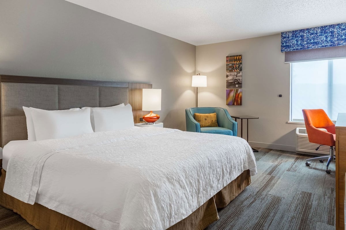 A hotel room with a large bed, a desk, a chair, a lamp, and a window.