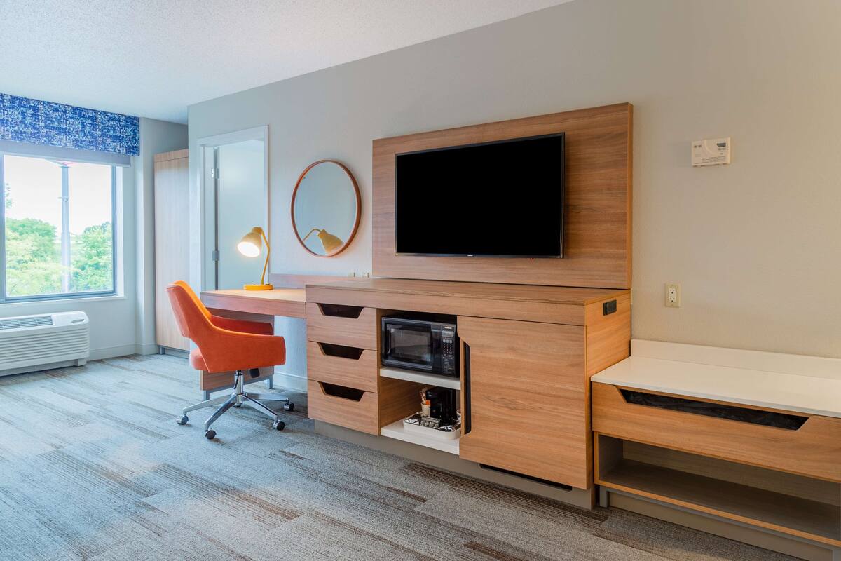 Habitación de hotel moderna con escritorio de madera, televisor de pantalla plana, un pequeño espejo y ventana con vistas a la vegetación.