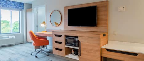 A modern hotel room with a wooden desk, a flat-screen TV, a small mirror, and a window with a view of greenery.
