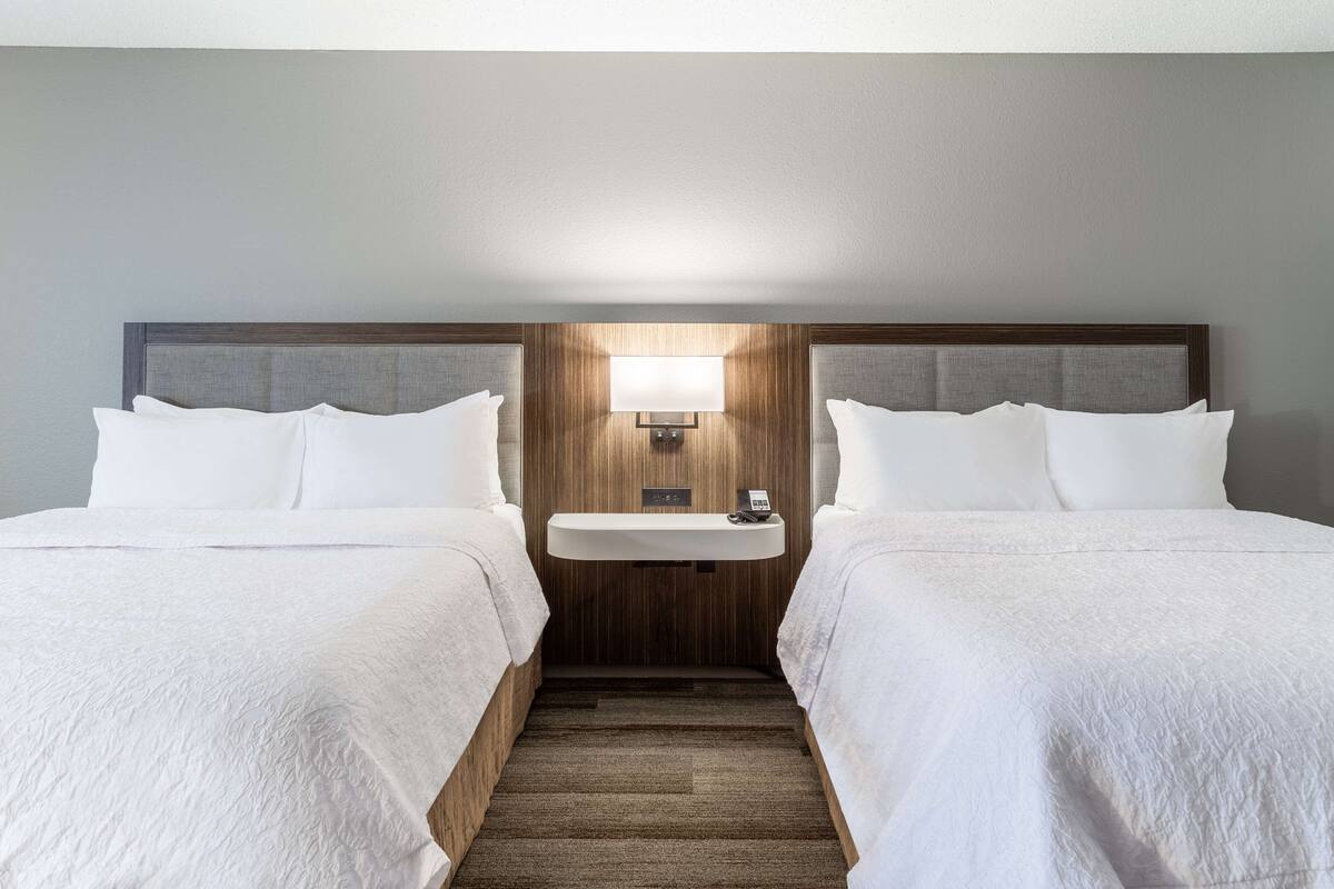 Habitación de hotel con dos camas, una mesita de noche pequeña con lámpara y un cabecero de madera.