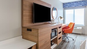 A modern hotel room with a wooden desk, a flat-screen TV, a mirror, an orange chair, and a large window with a view.