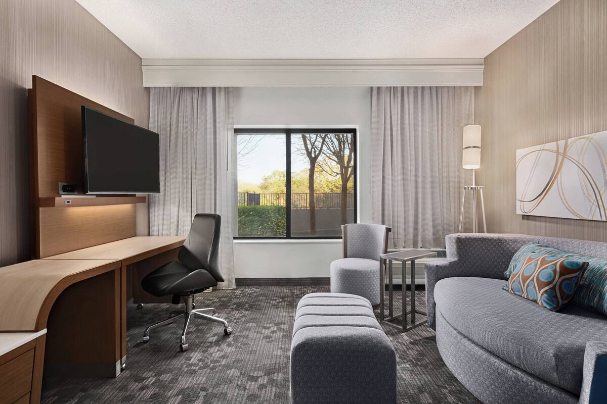 A hotel room with a desk, chair, sofa, ottoman, and a flat-screen TV.