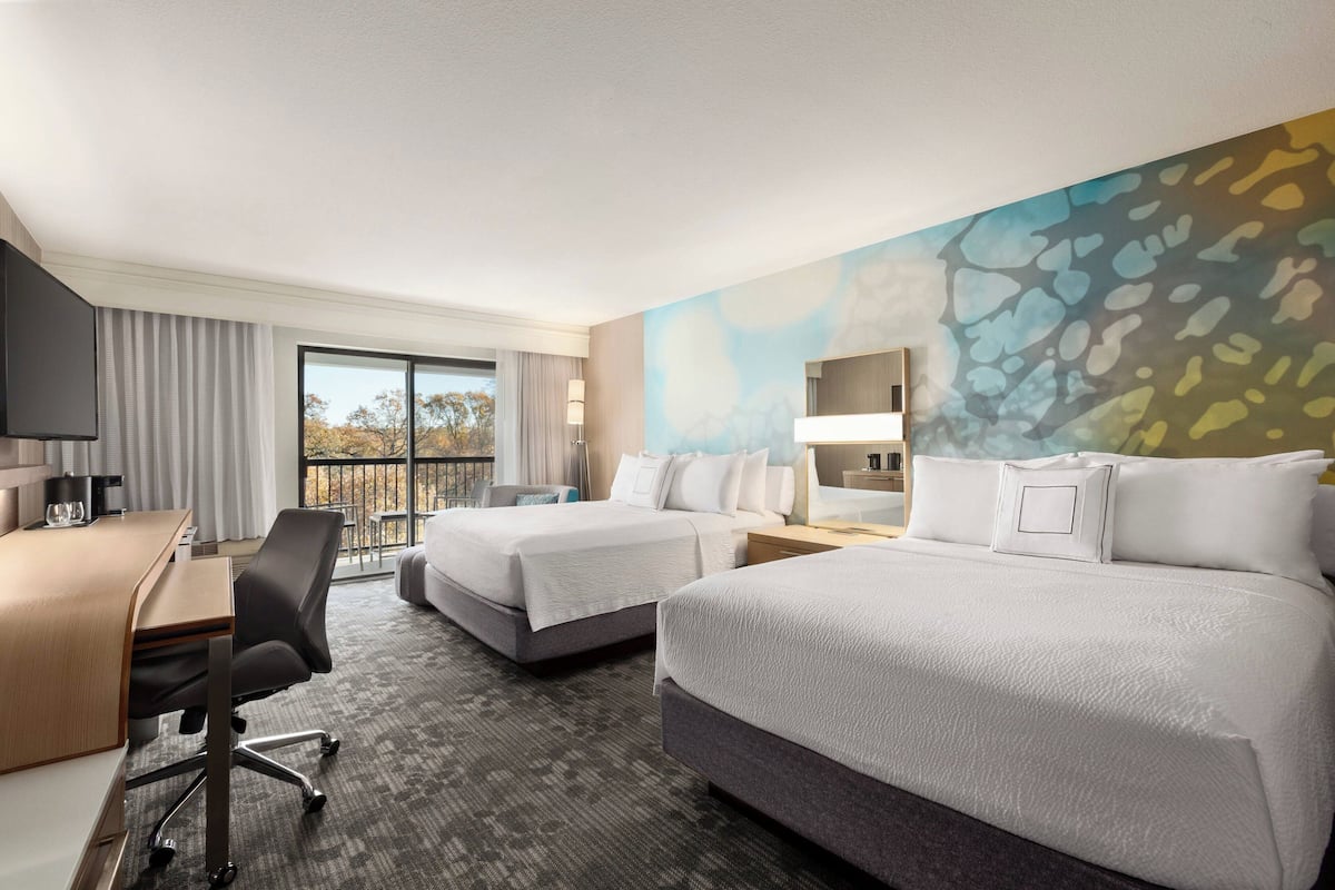 A hotel room with two beds, a desk, a chair, a TV, and a large window with a view of trees.