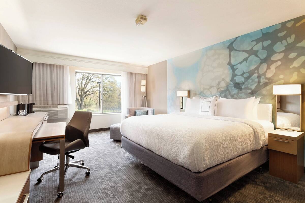 A hotel room with a large bed, a desk, and a chair. There is a window with curtains, a painting on the wall, and a lamp on the nightstand.