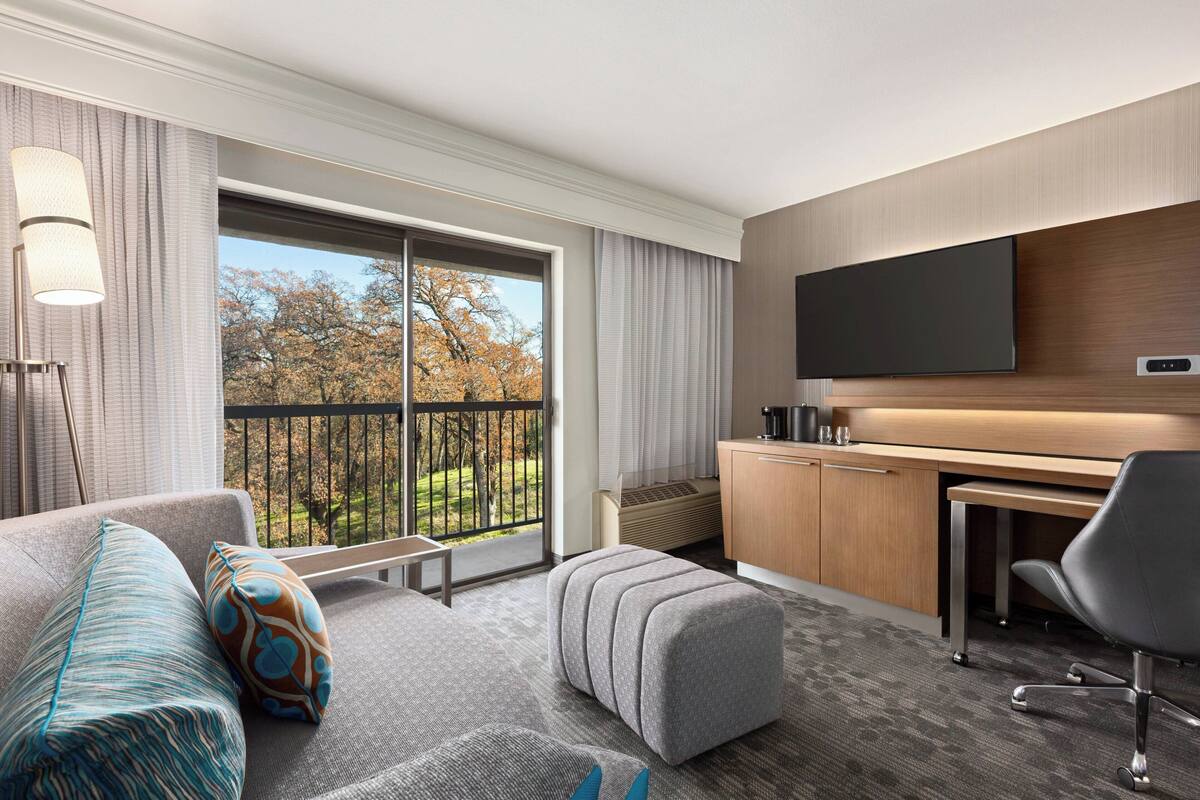 A modern hotel room with a large window, a flat-screen TV, a desk, and a comfortable seating area.
