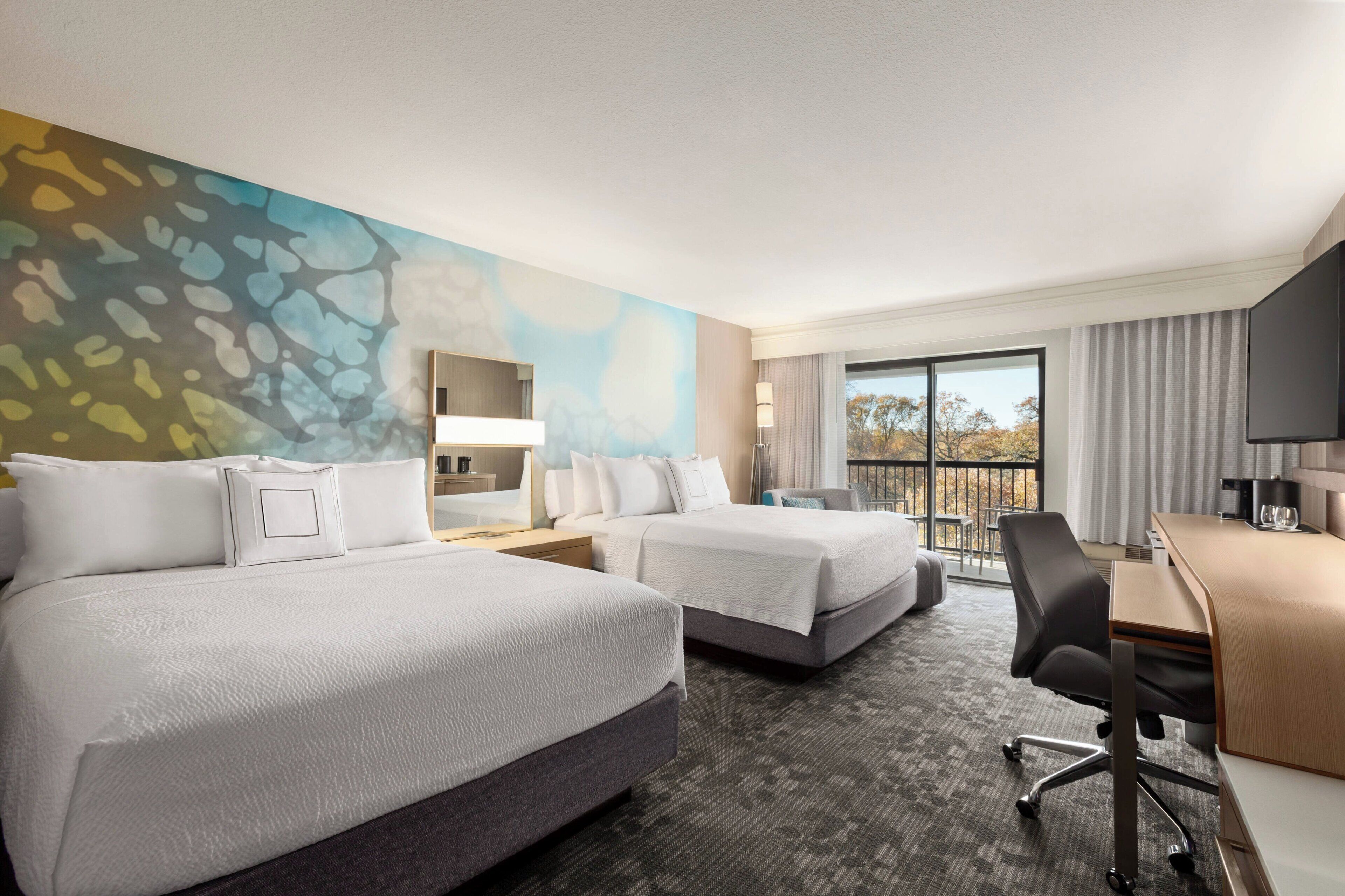 Ein Hotelzimmer mit zwei Betten, einem Schreibtisch, einem Sessel, einem Flachbildfernseher und einem Balkon mit Aussicht.