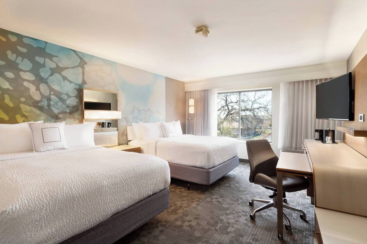 A hotel room with two beds, a desk, a chair, a TV, and a large window with a view.