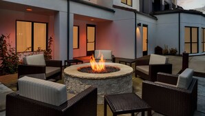 A circular stone fire pit surrounded by wicker furniture in an outdoor patio area with a building in the background.