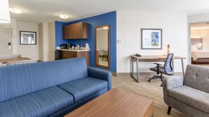A modern hotel room with a blue sofa, a wooden coffee table, a gray armchair, a desk with a chair, and a kitchenette area. - Holiday Inn Express & Suites Scottsburg by IHG (Scottsburg)