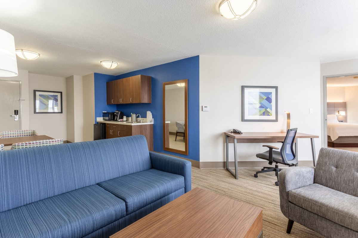 A modern hotel room with a blue sofa, a wooden coffee table, a gray armchair, a desk with a chair, and a kitchenette area.