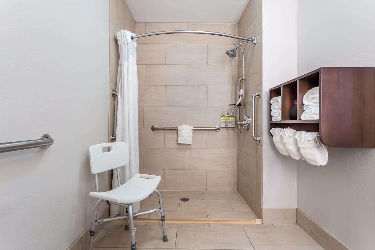 A wheelchair-accessible shower area with a grab bar, a white chair, and a shelf with towels.
