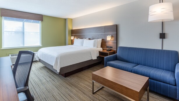 A hotel room with a bed, a blue sofa, a wooden coffee table, a desk with a chair, and a window with blinds. - Holiday Inn Express & Suites Scottsburg by IHG (Scottsburg)