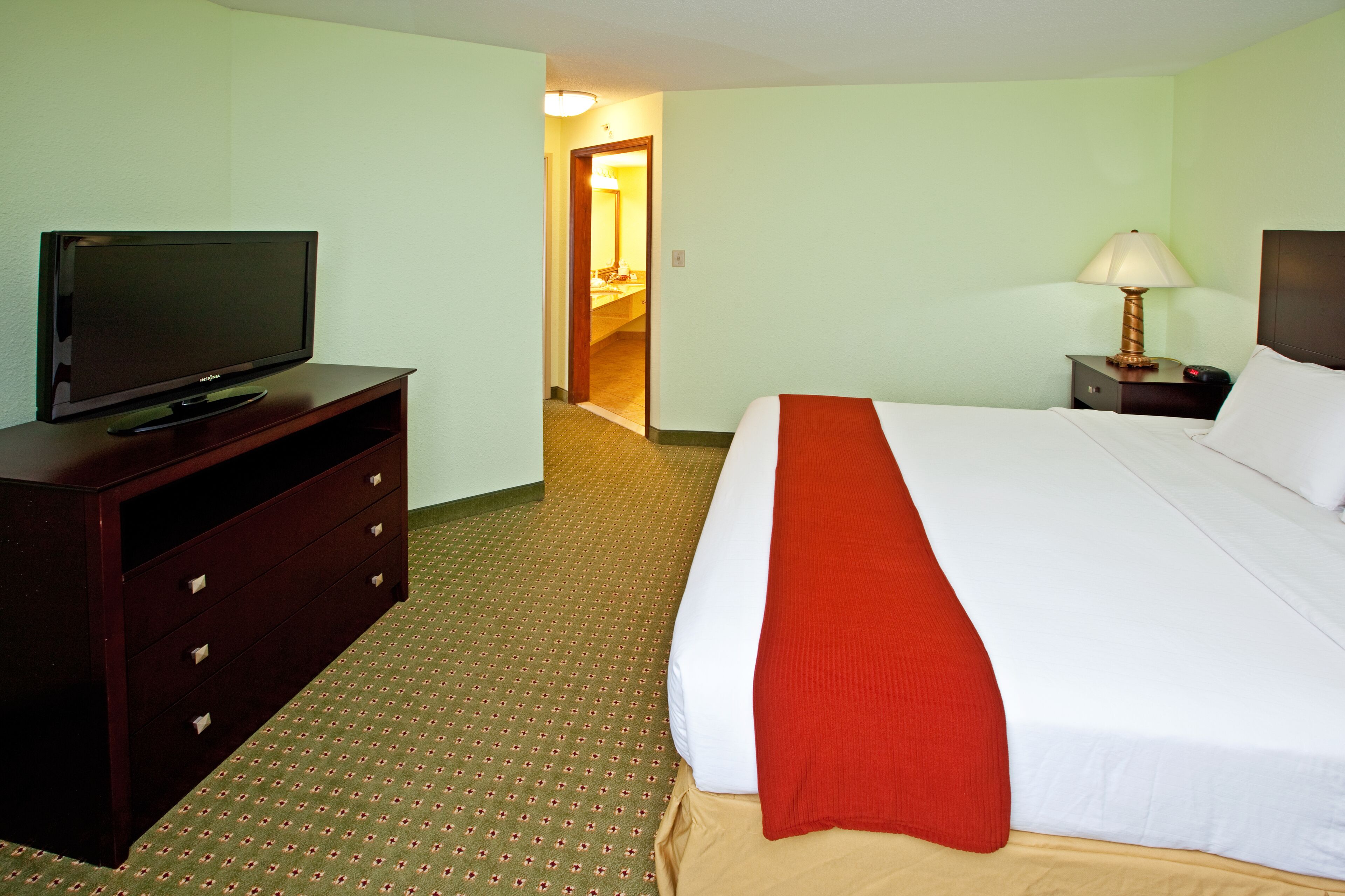 A hotel room with a bed, a television on a dresser, a lamp, and a door leading to a bathroom.