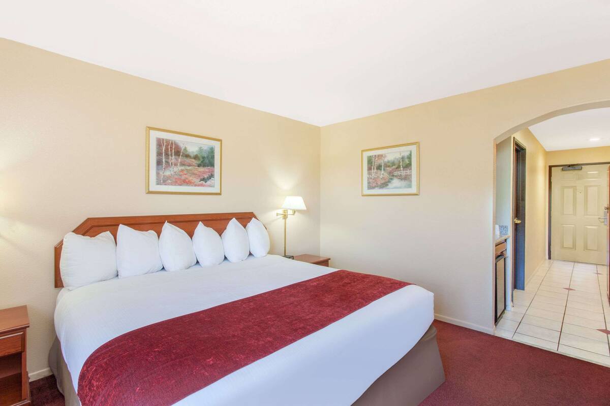 Hotelkamer met een groot bed, een nachtkastje, twee ingelijste foto's aan de muur, een lamp en een deur naar een andere kamer.