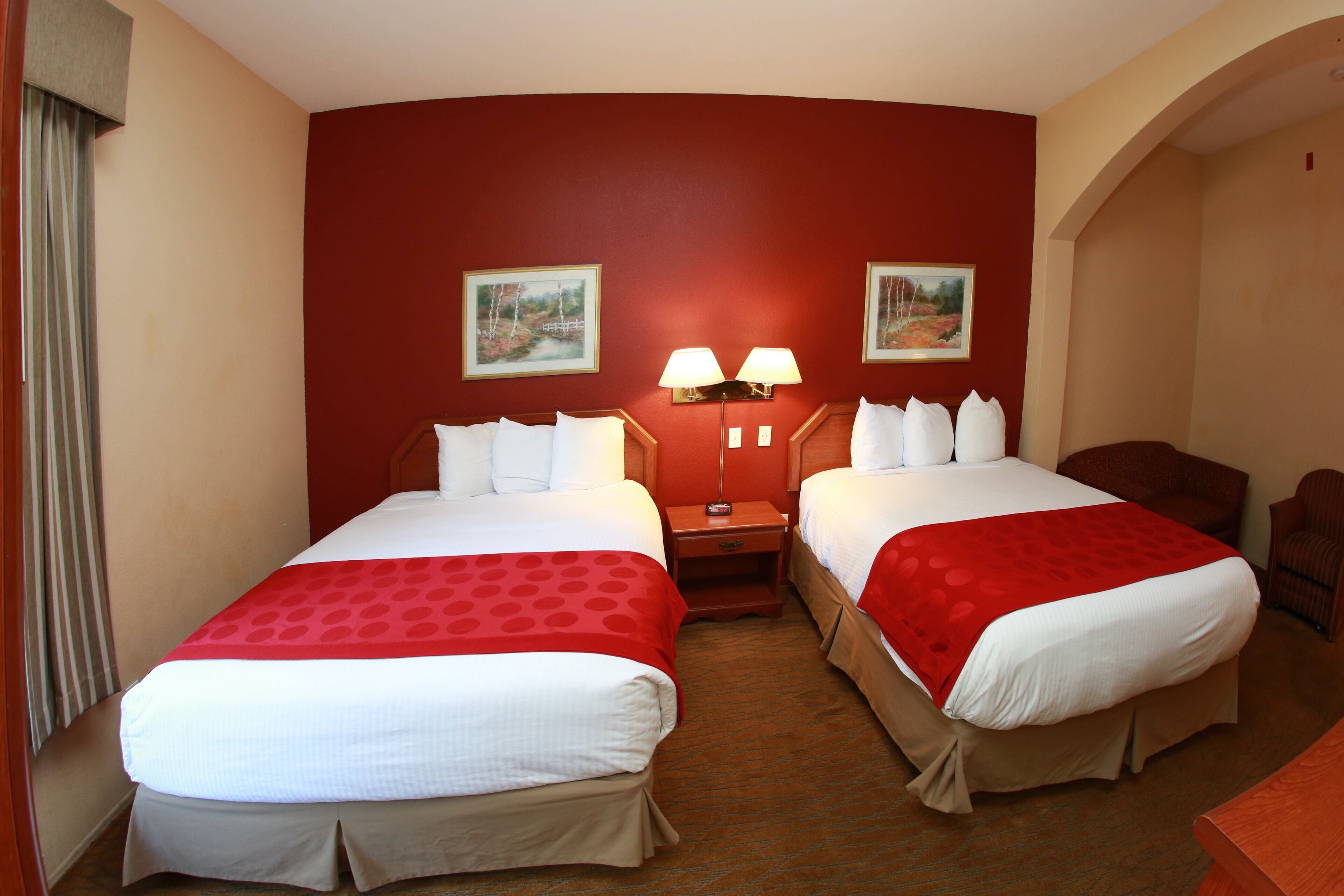 A hotel room with two beds, red and white bedding, a nightstand with lamps, and two wall-mounted paintings.
