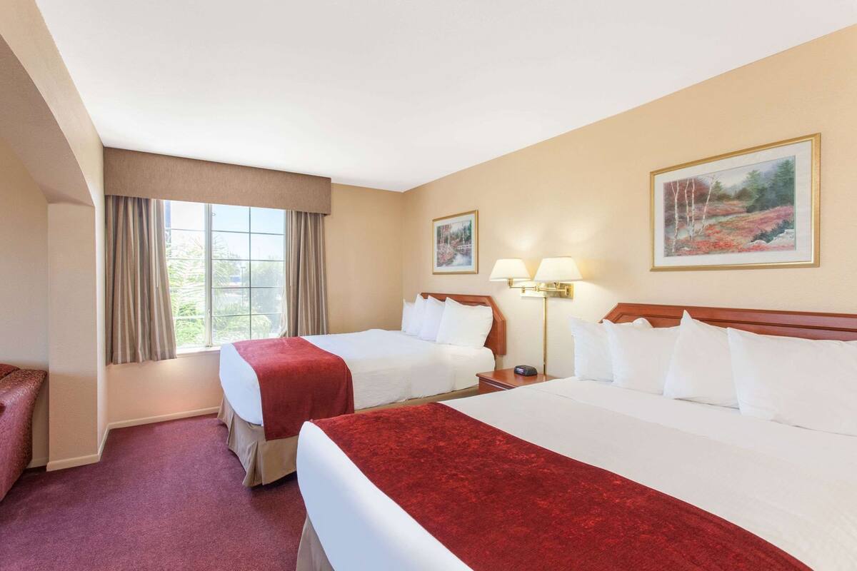 Een hotelkamer met twee bedden, een raam met gordijnen, een lamp en twee schilderijen aan de muur.