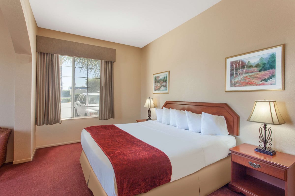 Een hotelkamer met een groot bed, een nachtkastje met een lamp, een raam met gordijnen en twee ingelijste foto's aan de muur.