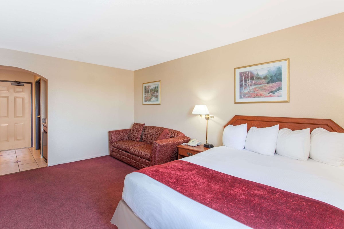 Een hotelkamer met een groot bed, een roodgekleurd tapijt, een bruine fauteuil, een nachtkastje met een lamp en twee ingelijste foto's aan de muur.