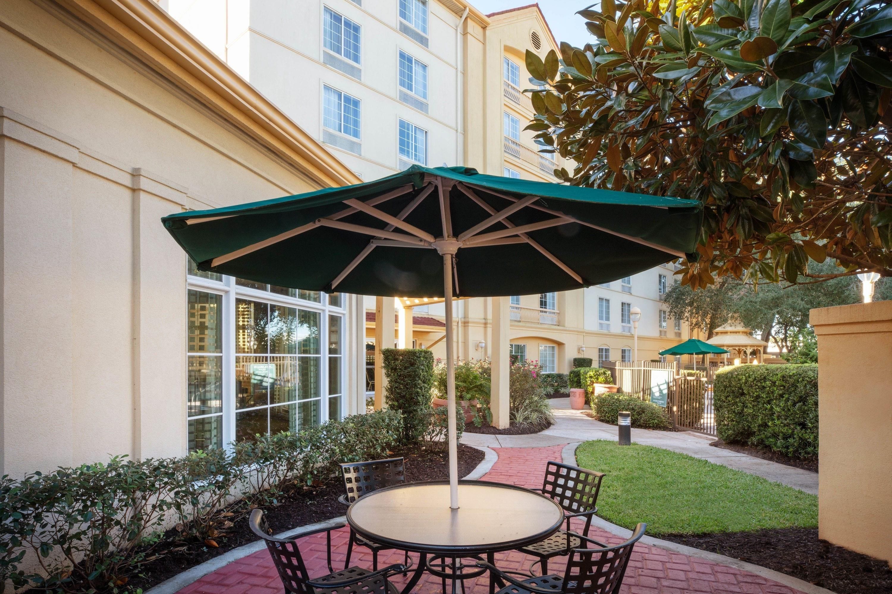 An outdoor patio with a round table and chairs, a green umbrella, and a view of a building with multiple windows.