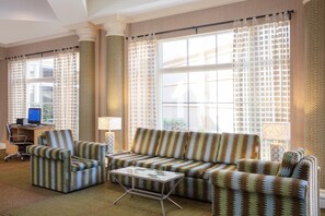 A hotel room with a sofa, armchair, and a small table. There is a desk with a computer and a lamp. The room has large windows with patterned curtains.