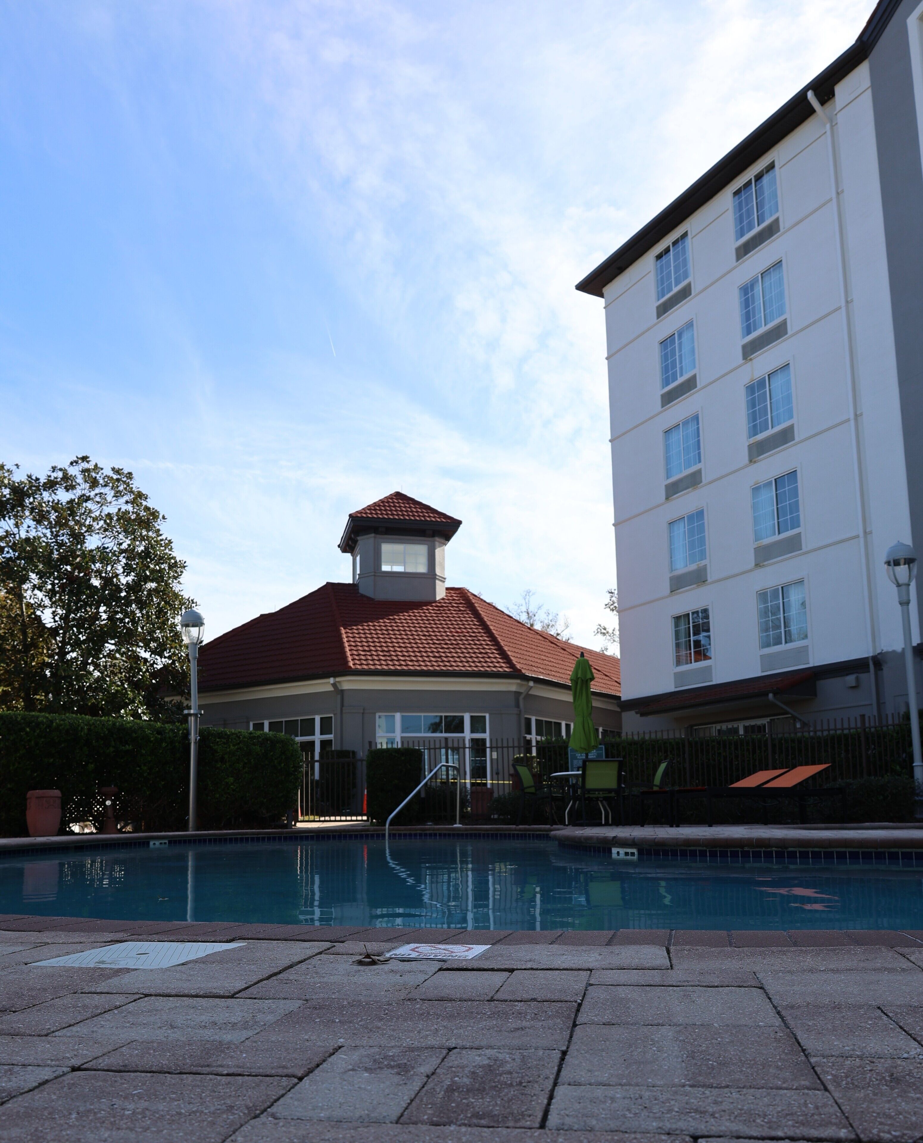 Une piscine avec une terrasse carrelée, un bâtiment doté d’un toit rouge et d’une tour, et une clôture avec un portail.