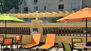 Un espacio de estar al aire libre con sillones y mesas, una pérgola con sombrillas, y un edificio con varias ventanas al fondo.