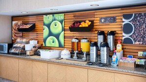 Zona de recepción de un hotel con letrero de cafetería, una exhibición de frutas y varios dispensadores de café y té.
