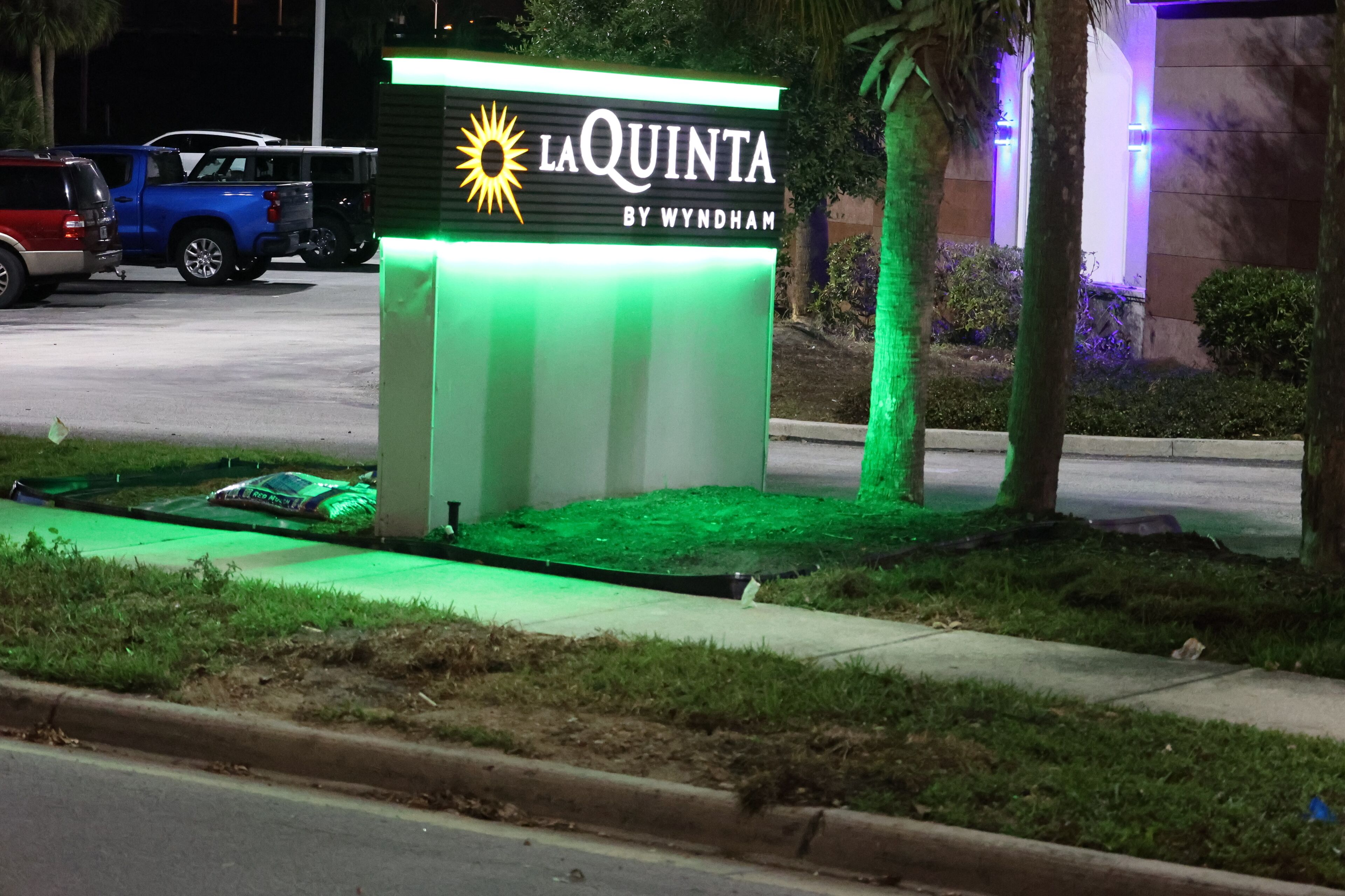 A sign for "La Quinta by Wyndham" with a sun logo, surrounded by greenery and parked vehicles.