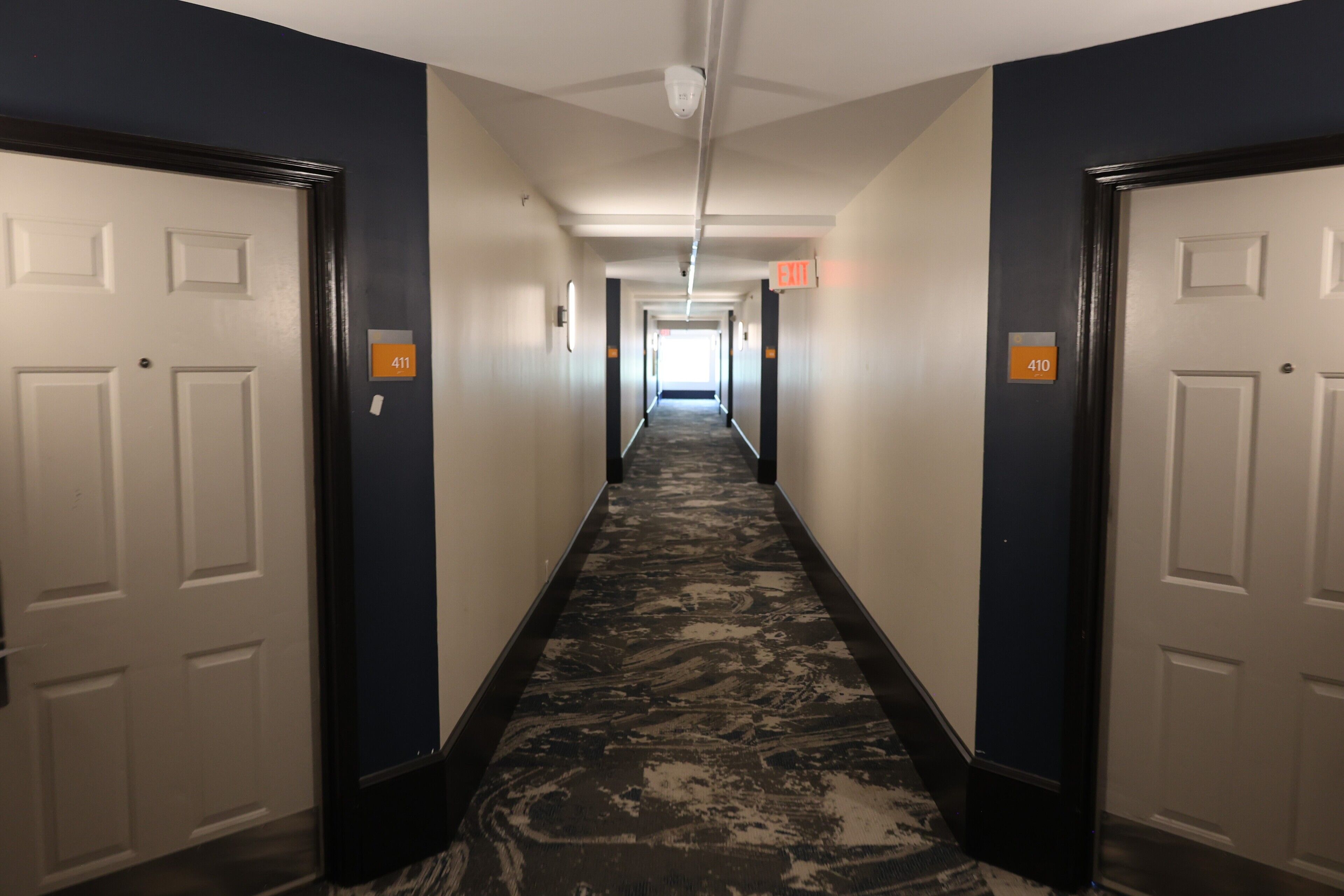 A hotel hallway with a carpeted floor, white doors, and orange exit signs.