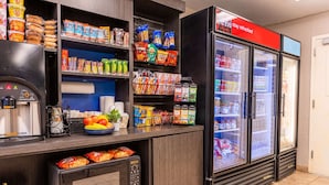 Eine Selbstbedienungs-Speiseausgabe mit Kaffeeautomat, Mikrowelle und Kühlschrank, gefüllt mit Snacks und Getränken.