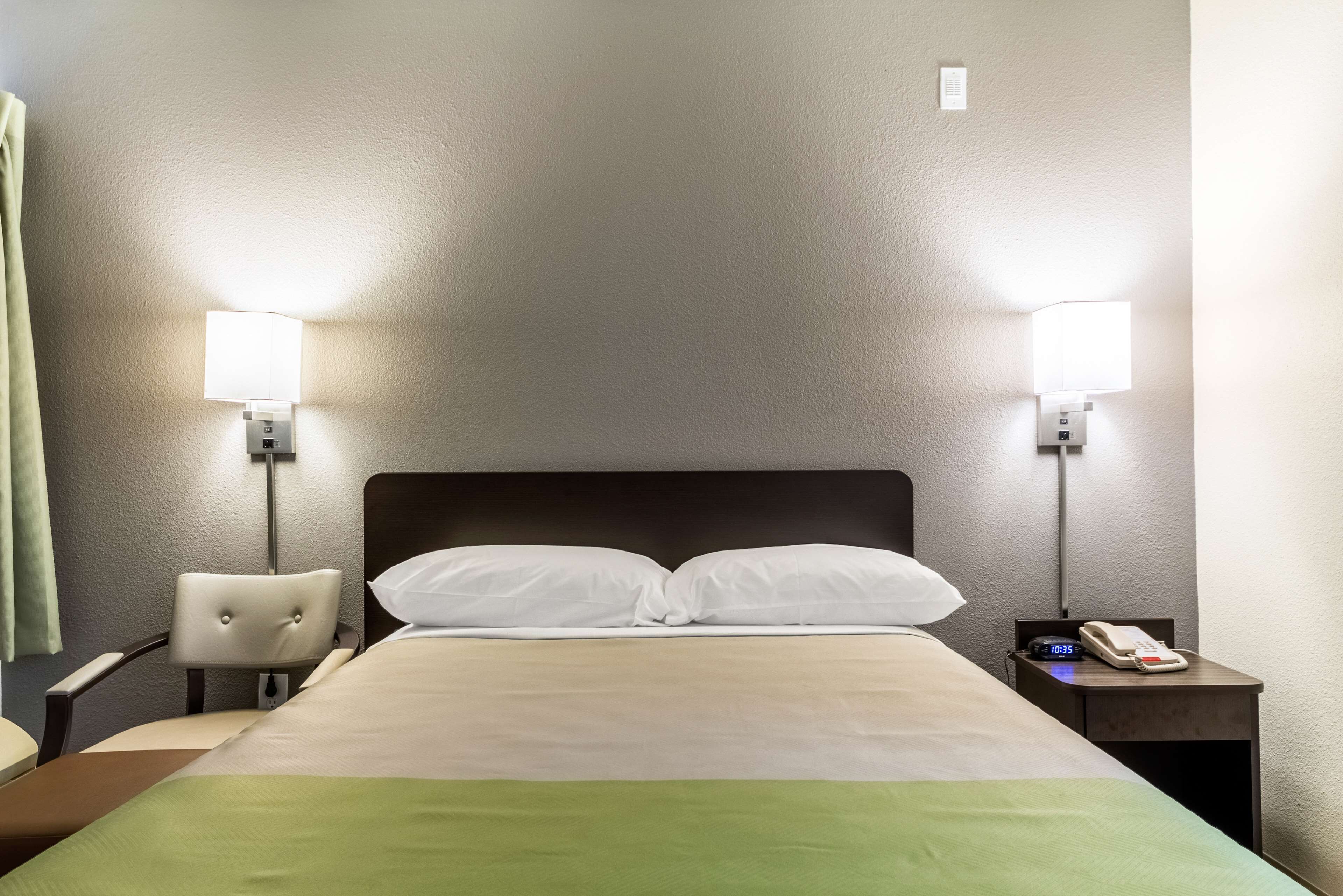 Ein Hotelzimmer mit einem Bett, zwei Nachttischlampen, einem Sessel, einem Nachttisch mit Telefon und einer Wanduhr.