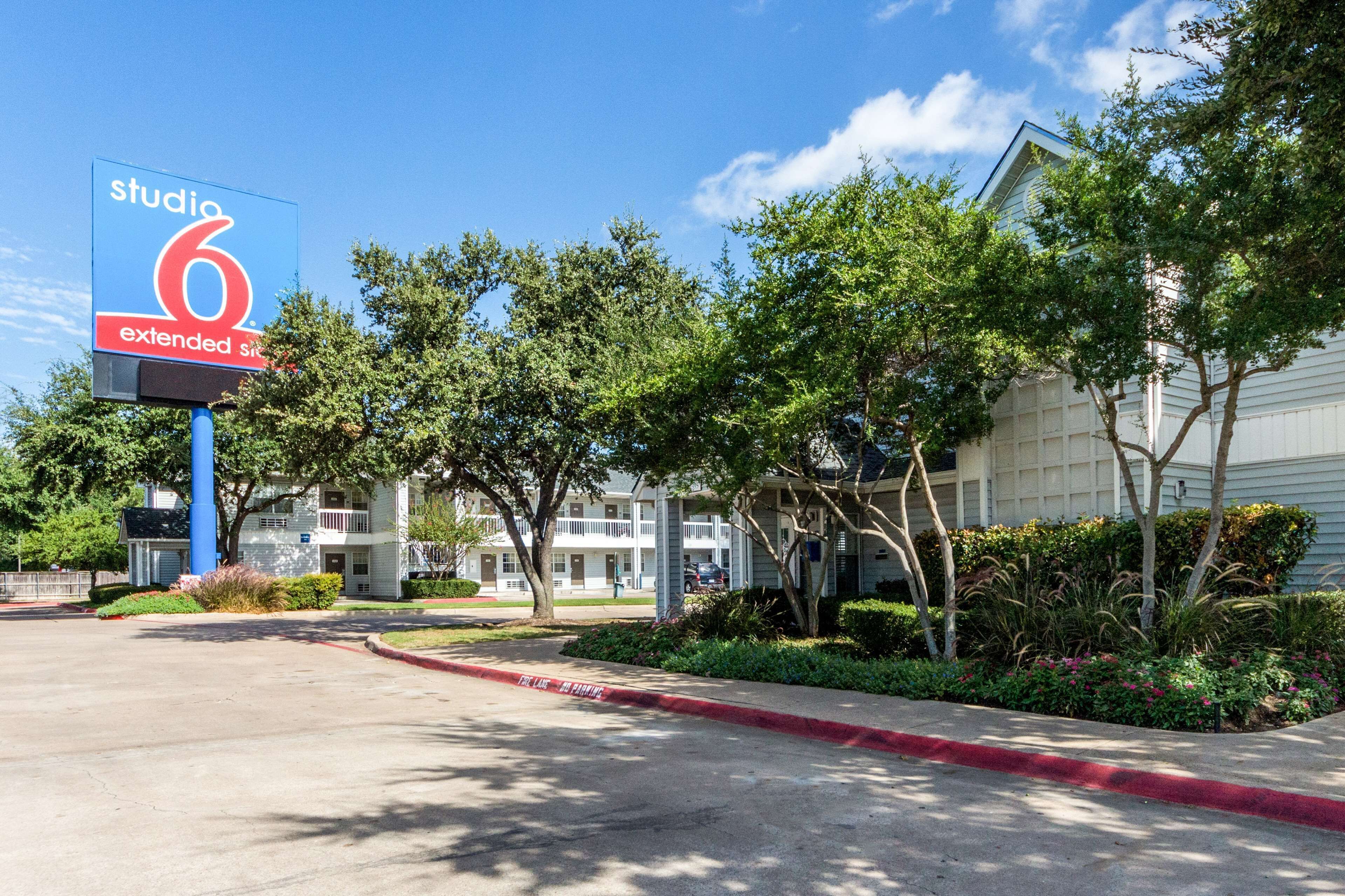 A hotel named 'Studio 6 Extended Stay' with a parking lot, trees, and a building with a white facade.