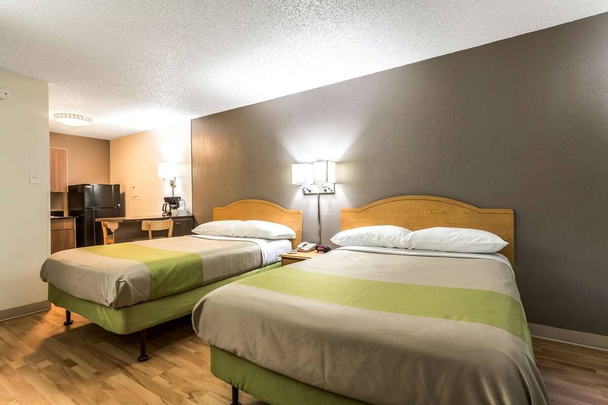 Ein Hotelzimmer mit zwei Einzelbetten, einem Holzkopfteil, einem Schreibtisch mit Stuhl, einem kleinen Tisch, einer Lampe und einem Fernseher.