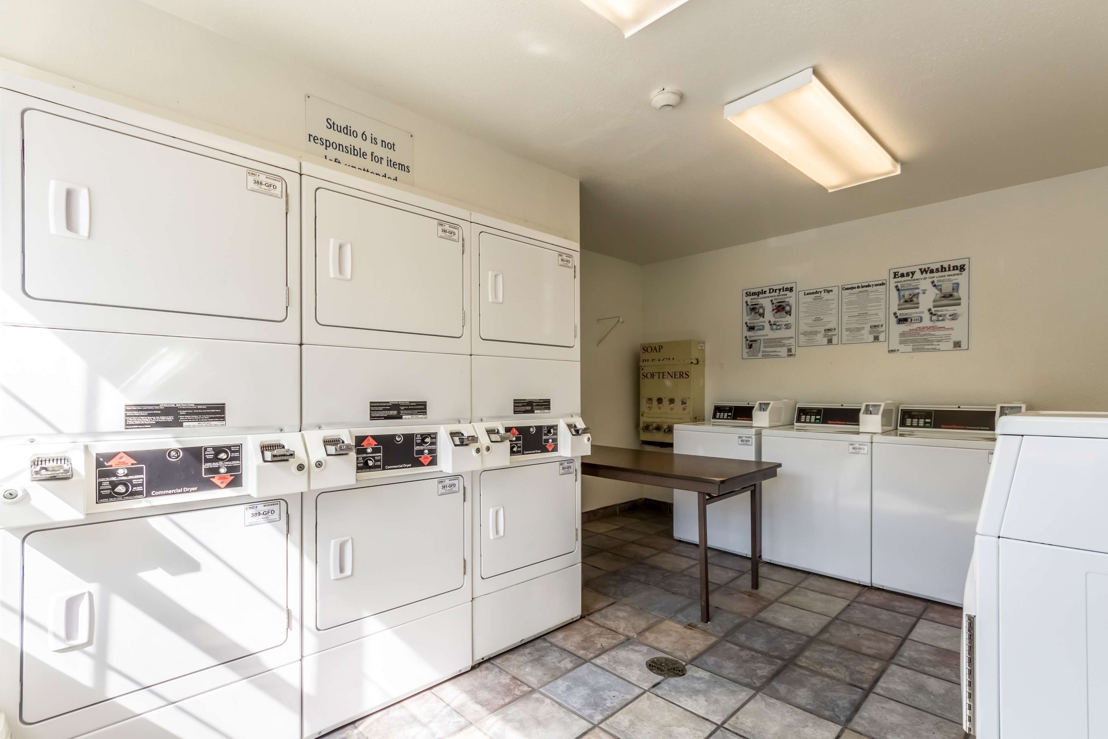 A laundry facility with multiple washing machines and dryers, a table, and informational posters on the wall.